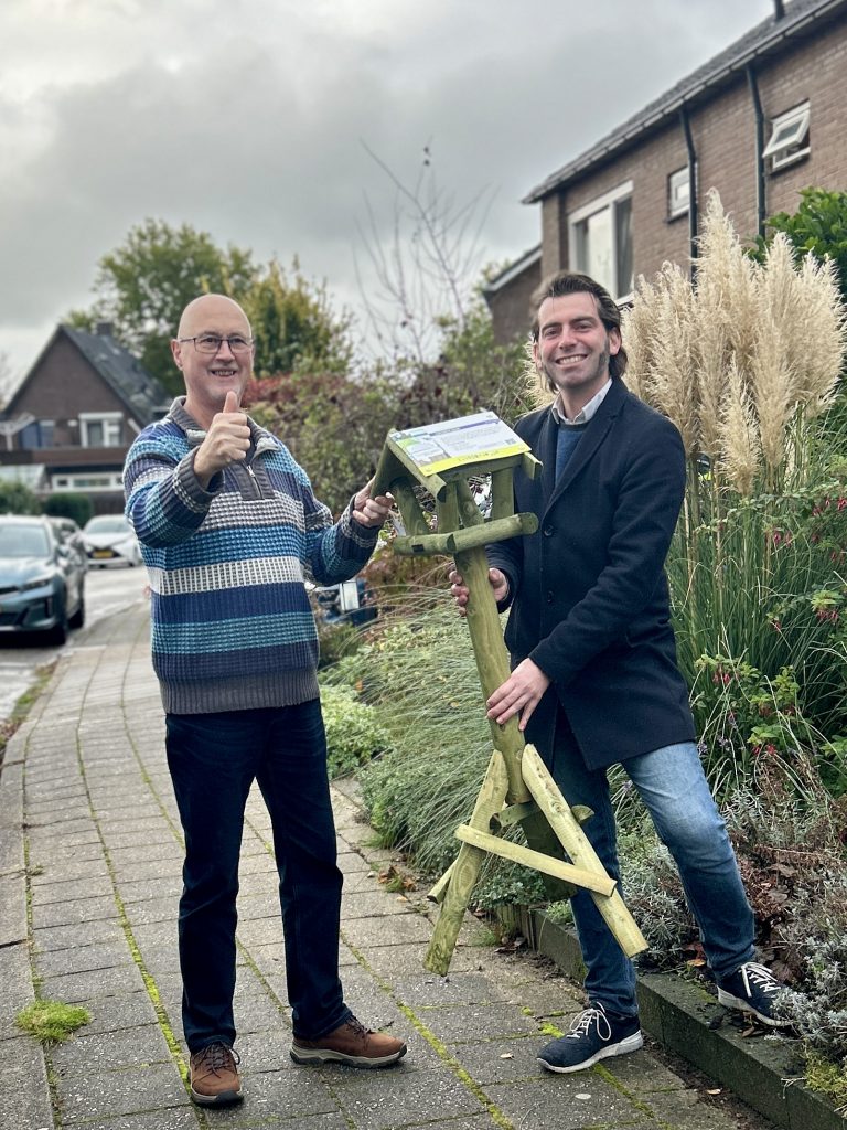 Afbeelding met wethouder Paul Hofman van gemeente Rheden, die een vogelvoederhuisje uitreikt aan een inwoner van Dieren-West vanwege het nemen van een duurzame maatregel